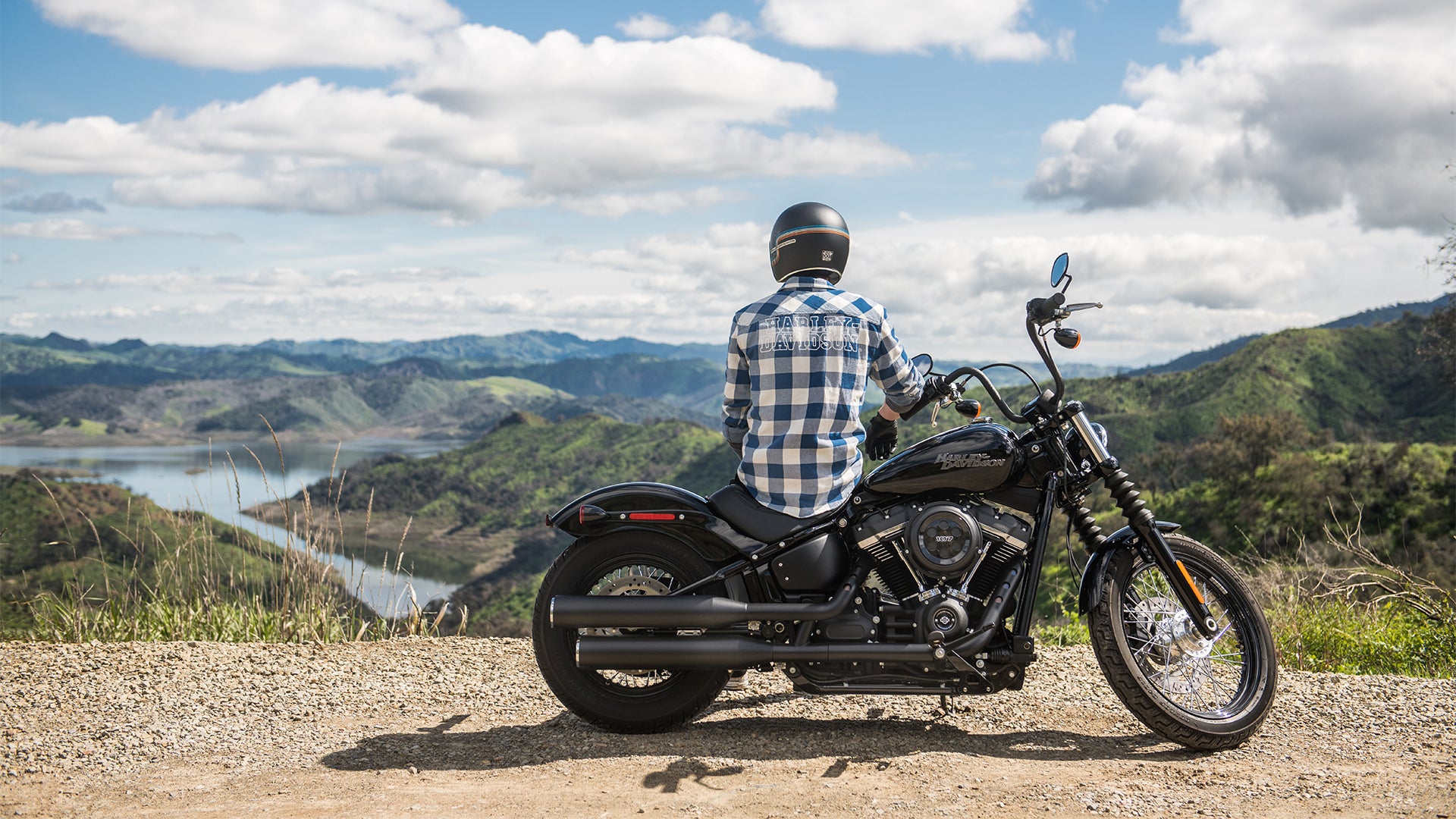 A man sitting on a black motorcycle parked on the side of a dirt road overlooking some green mountains and a lake.