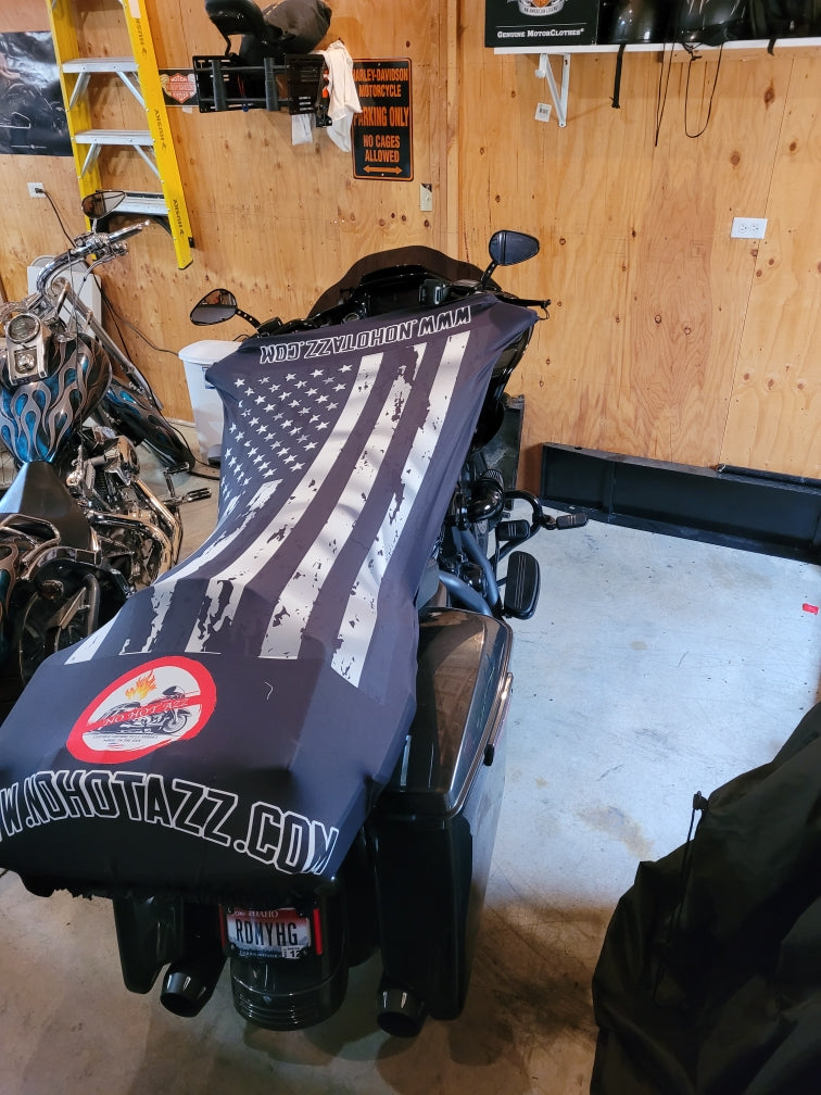 A rear view of a black No Hot Azz motorcycle seat shade sun cover on a motorcycle in a garage featuring a black and white american flag.