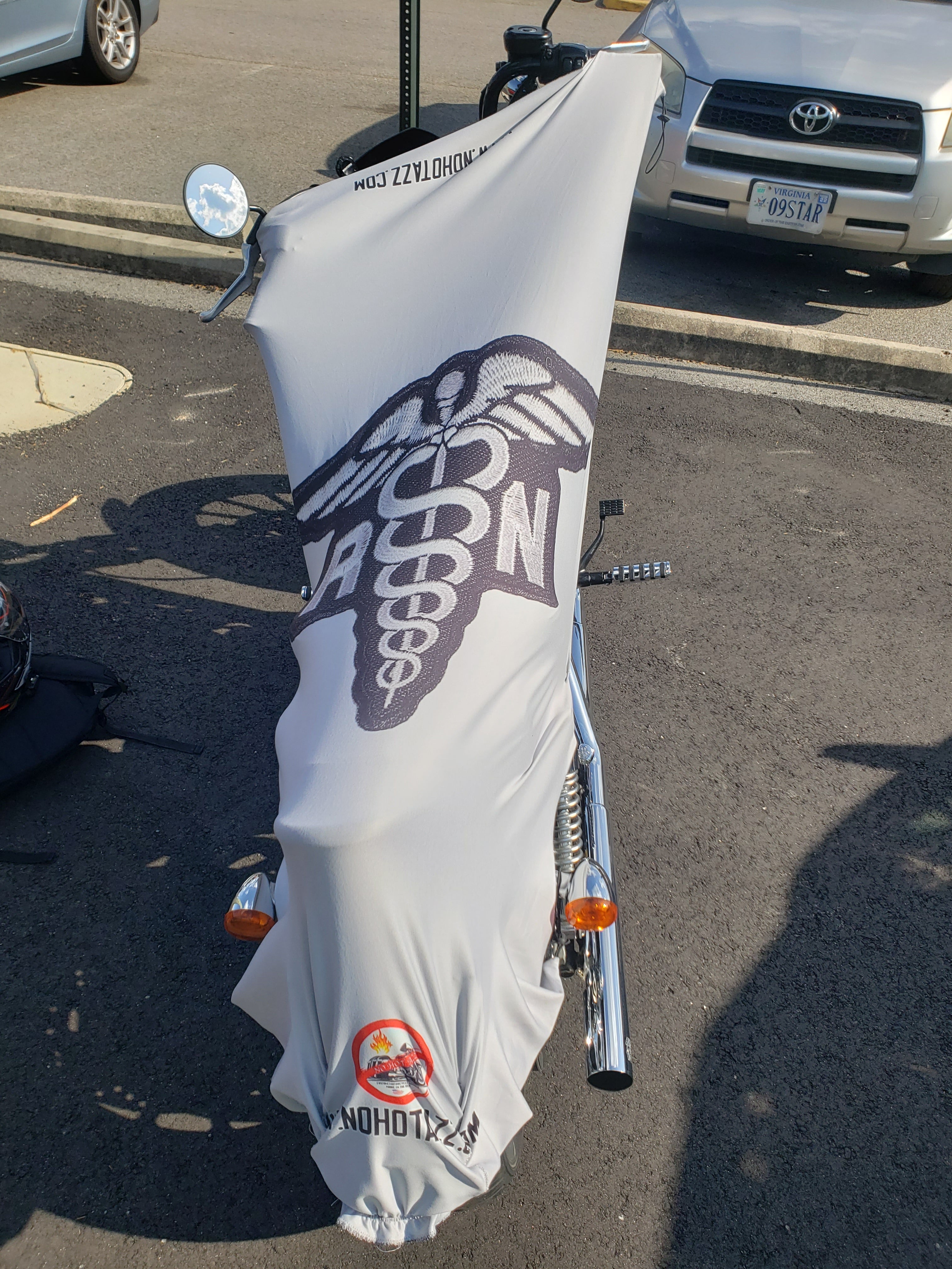 A rear view of a white No Hot Azz motorcycle seat shade sun cover on a motorcycle in a parking lot featuring a registered nurse patch.