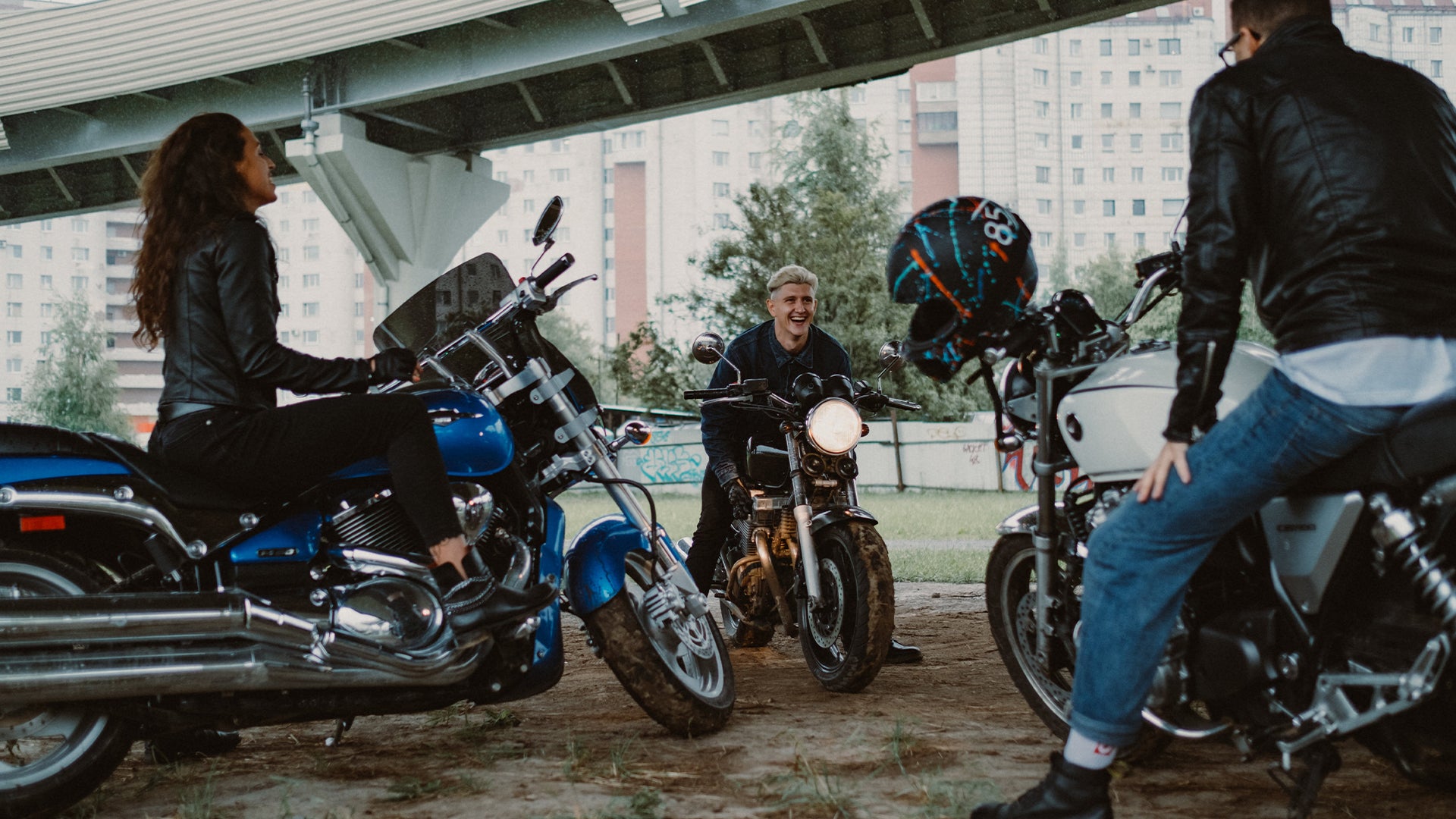 Three smiling motorcycle riders sitting on their motorcycles under an overpass.